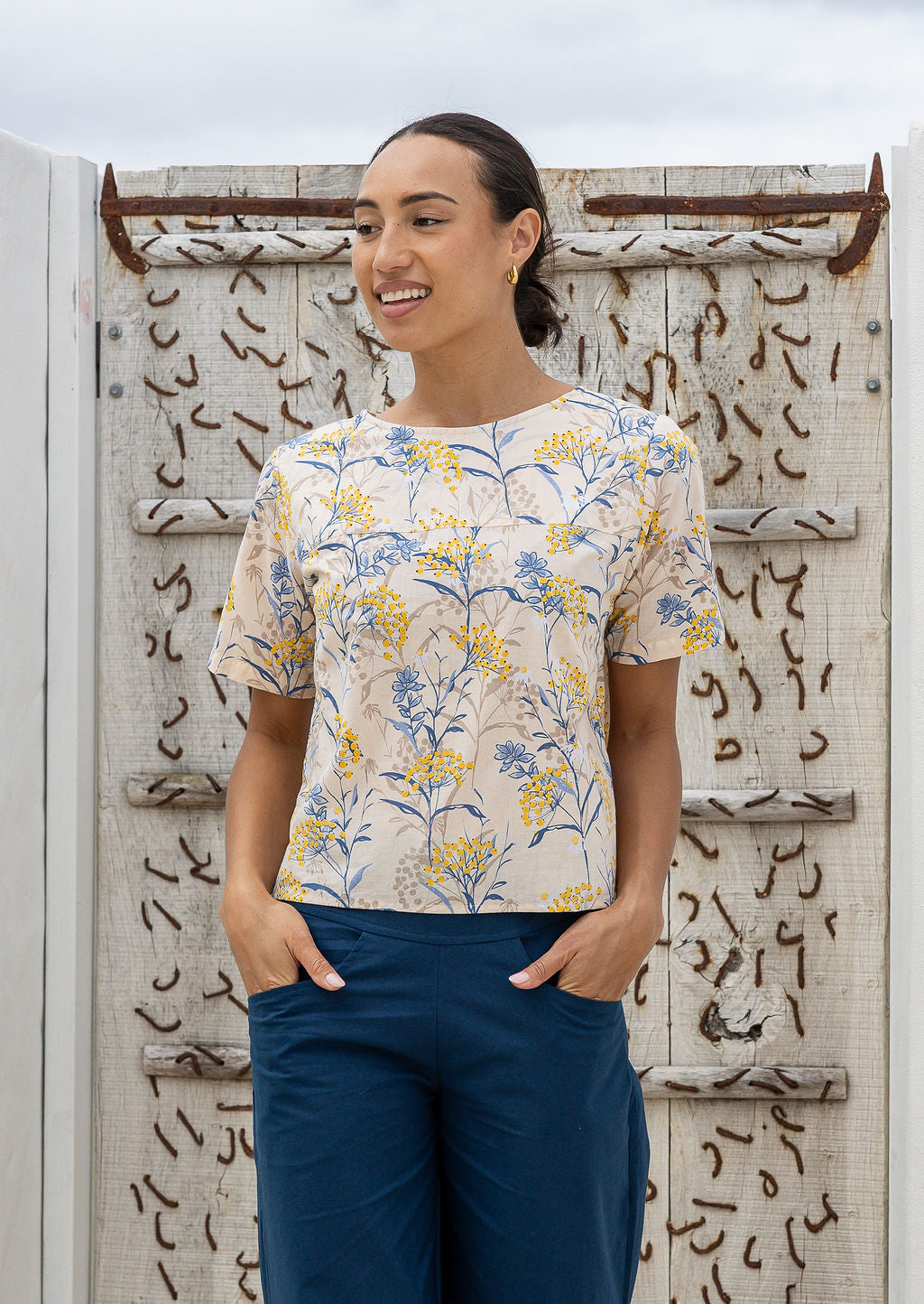 Woman wearing a loose fit neutral based floral with blue pants against a white wall.