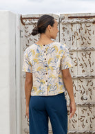 Woman wearing a floral blouse and dark pants standing against a textured wall.