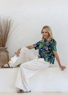 Woman sits on a white ledge in a bright, minimalist room, wearing a short-sleeve 100% cotton top paired with white plain pants