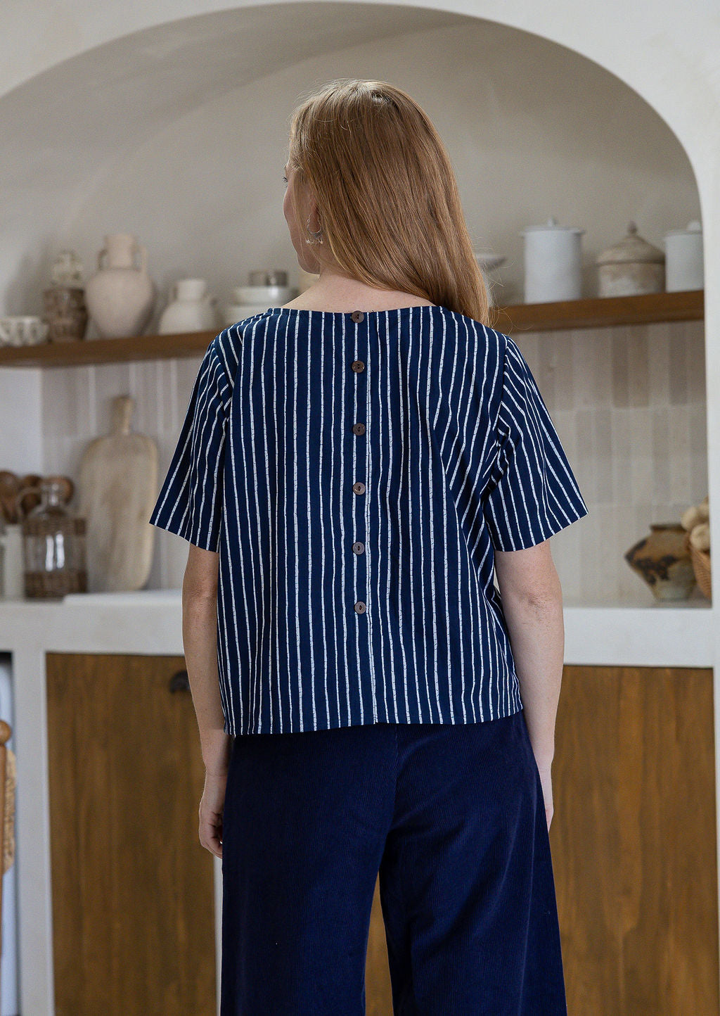Back view of woman wearing relaxed fit navy and white striped cotton top with decorative buttons down the centre back