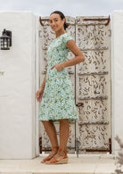 Model with one hand in pocket wears a knee length A line dress with cap sleeves in sage green based floral print