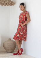 Woman leans on wall posing in ared based floral sundress with round neckline, cap sleeves, A line skirt and pockets