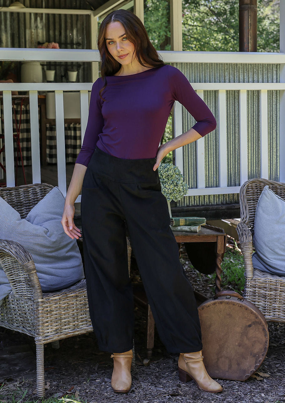 Woman in a purple top and black cotton corduroy pants standing in front of a porc and wicker chairs