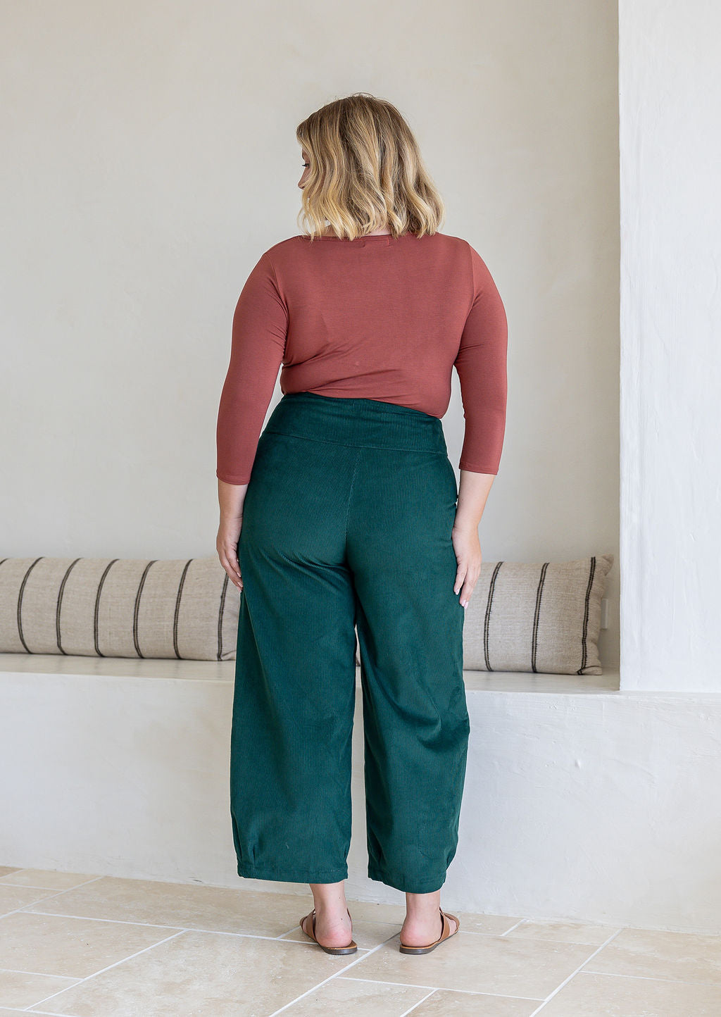Model standing backwards wears corduroy wide leg pants with side zipper, pockets and small pleats at ankles.
