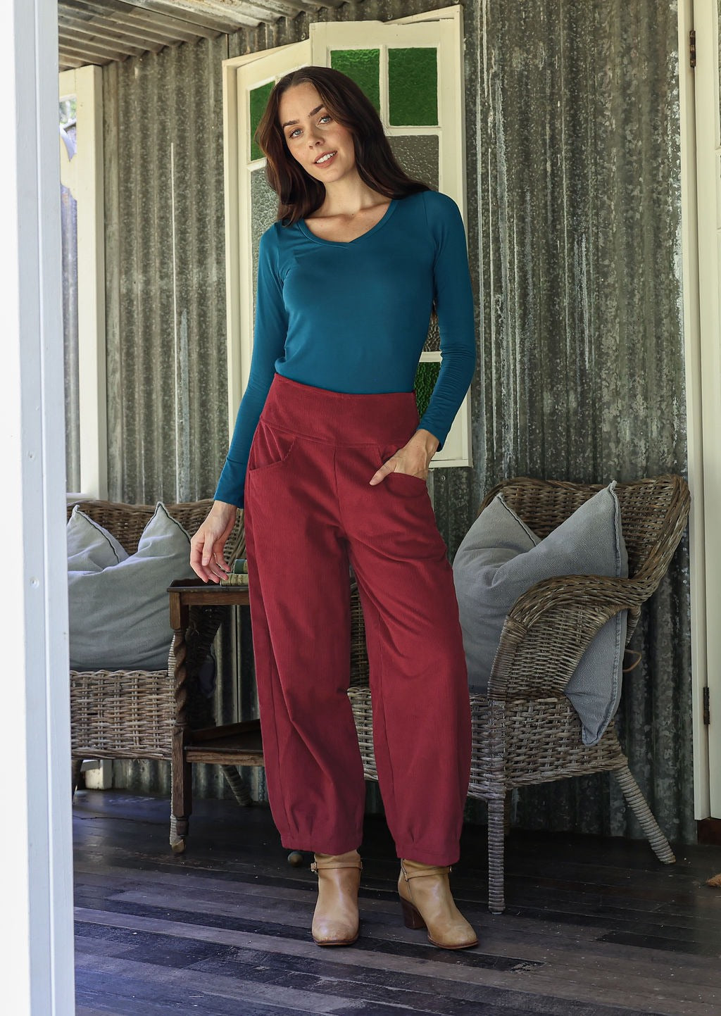 Woman in teal top and red corduroy pants standing on a wooden deck with wicker chairs and cushions.