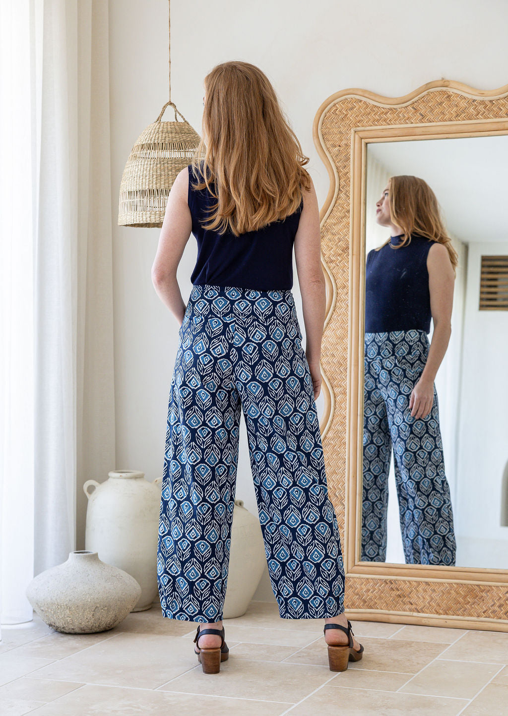 Model standing backwards wears 100% cotton wide leg pants with side zipper, pockets and small pleats at ankles.
