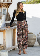 Woman posing in 100% cotton wide-leg black based floral printed pants with wide flat waistband, side zipper and small pleats at the ankles.