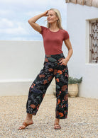 Woman posing in 100% cotton wide-leg black botanical printed pants with wide flat waistband, side zipper and small pleats at the ankles.