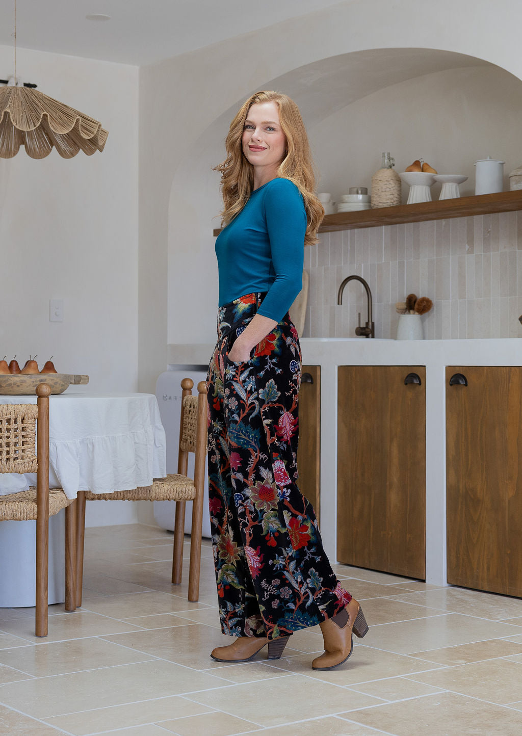Woman standing sideways wearing a black based multi-colour floral velvet wide leg pants with basic blue top