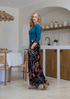 Woman standing sideways wearing a black based multi-colour floral velvet wide leg pants with basic blue top