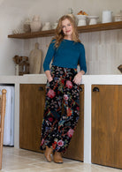 Woman standing in a kitchen wearing a blue top and floral pants.