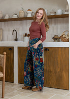 Woman wearing a basic top with teal based multi-colour floral velvet wide leg pants with hands in pockets and a side zip