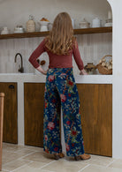 Woman standing backwards wearing a teal based multi-colour floral velvet wide leg pants with wide waistband 