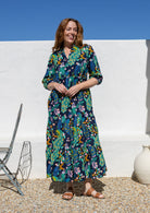 A woman wearing a 3/4 sleeved maxi dress with a vibrant multicolour floral and peacock print on a dark navy background, standing outdoors against a white wall under a clear blue sky.