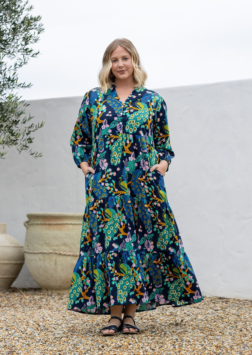 A woman wearing a 3/4 sleeved maxi dress with a floral and peacock print with both hand in pockets