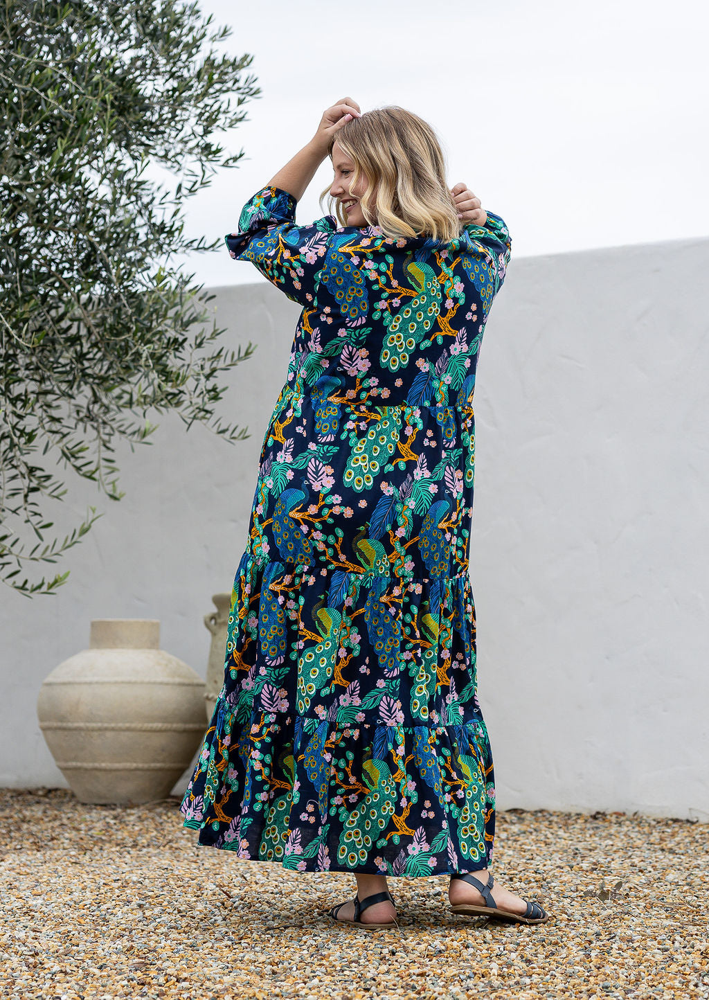 A woman stands backwards wearing a 3/4 sleeved cotton maxi dress with a colourful floral and peacock print. 