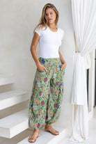 Woman wearing a white top and moss green floral pants with wide elastic waist and ankles standing in front of a white wall 