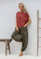 Woman standing in front of white wall wears loose fit cotton pants in olive green with deep pockets and elasticated ankles