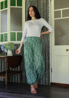 Woman wearing a white top and sage green floral pants with wide elastic waist and ankles standing in a rustic room