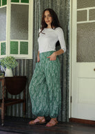 Woman wearing a white rayon top and sage green based floral pants with hands in deep pockets.