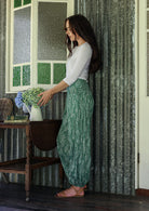Woman holding flowers wearing a 100% cotton floral green harem pants with flat wide waistband in front and elastic at the back.