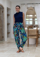 A woman modeling a sleeveless navy blue mock-neck top and vibrant, multicolored paisley-patterned cotton harem pants in a bright, modern indoor setting with a wicker pendant light and wooden dining chair.