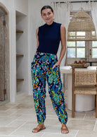 A woman wearing a sleeveless navy top and high-waisted, wide-leg cotton harem pants with a vibrant multicolor floral and bird pattern. She has her hands in the pockets. 