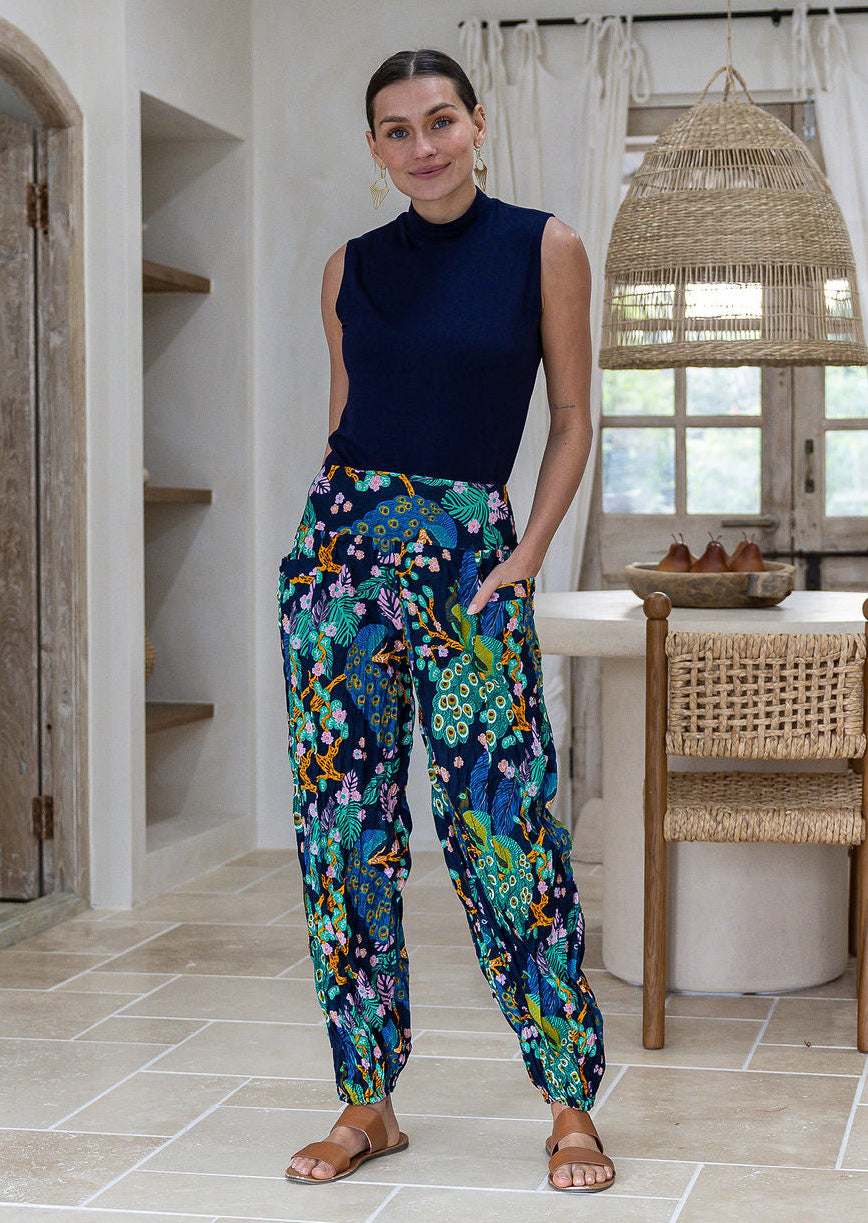 A woman wearing a sleeveless navy top and high-waisted, wide-leg cotton harem pants with a vibrant multicolor floral and bird pattern. She has her hands in the pockets. 