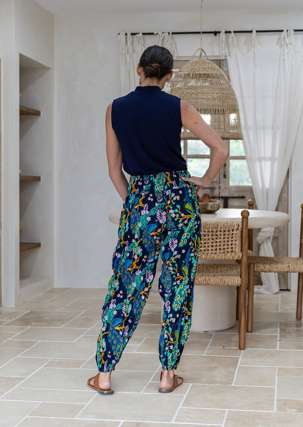 Back view of woman wearing cotton harem pants with a peacock print and elastic ruched back, paired with a sleeveless navy top. 