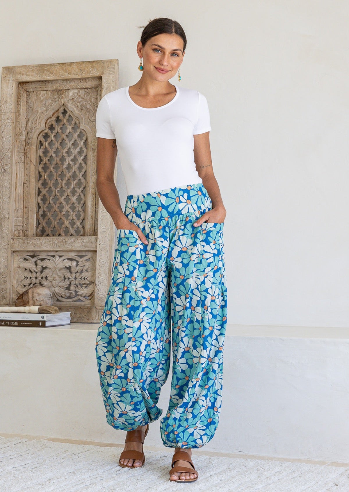 Woman poses in cotton harem pants in blue and aqua daisy print, with hands in pockets, paired with a white top