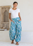 Woman poses in cotton harem pants in blue and aqua daisy print, with hands in pockets, paired with a white top