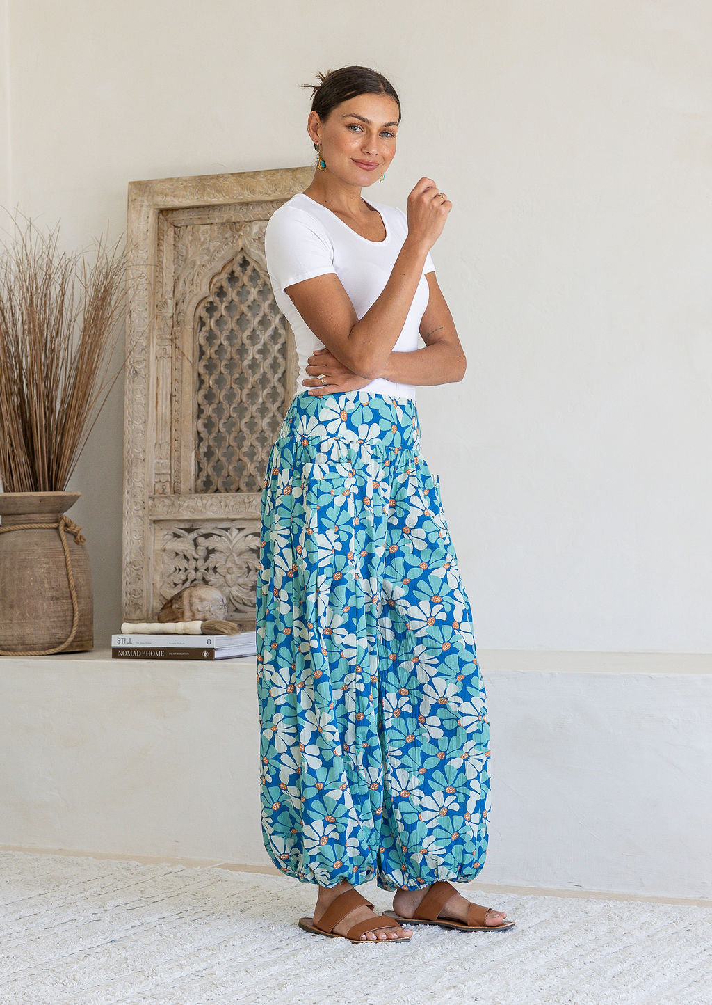 A woman modelling a pair of loose, cotton harem pants with a blue daisy print, paired with a white t-shirt and tan sandals, standing in a bright, minimalist room.
