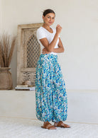 A woman modelling a pair of loose, cotton harem pants with a blue daisy print, paired with a white t-shirt and tan sandals, standing in a bright, minimalist room.
