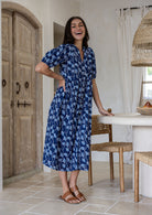 A woman wearing a navy blue based floral midi dress stands in a  kitchen setting. The dress features puff sleeves and side pockets.