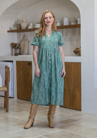 Woman standing on in a sage green based  floral cotton midi length dress standing in a kitchen