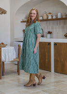 Woman standing side on in a sage green floral cotton midi length dress standing in a kitchen.