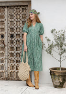 Woman in a sage green based floral print dress standing next to a wooden door and potted plant