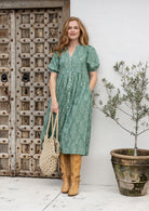 Woman in a sage green based floraldress standing in front of a wooden door and potted plant.