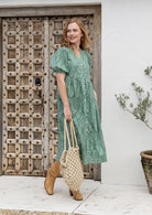Woman in a sage green based floral dress holding a woven bag in front of a wooden door.