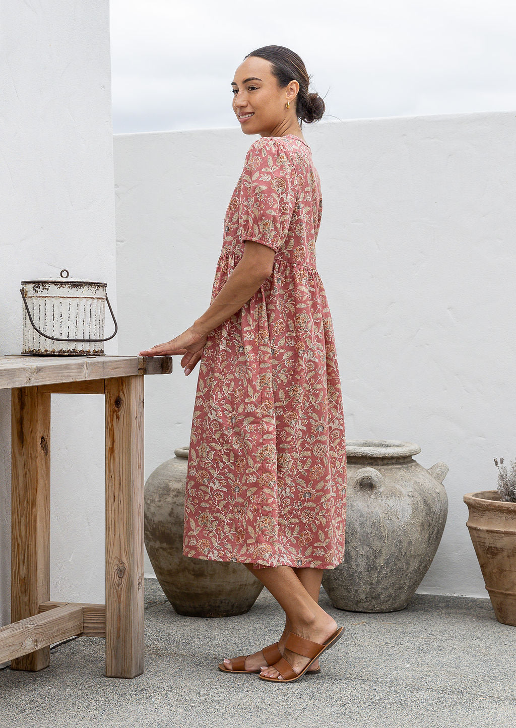 Woman standing side on in a pink floral cotton midi length dress standing next to a wooden table with decorative pots in the background.