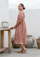 Woman standing side on in a pink floral cotton midi length dress standing next to a wooden table with decorative pots in the background.