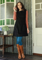 Woman wearing a black cotton corduroy sleeveless tunic paired with dusty pink top, blue leggings and tan boots