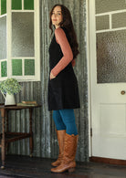 Woman standing side on in a black cotton corduroy sleeveless tunic with hands in front pockets