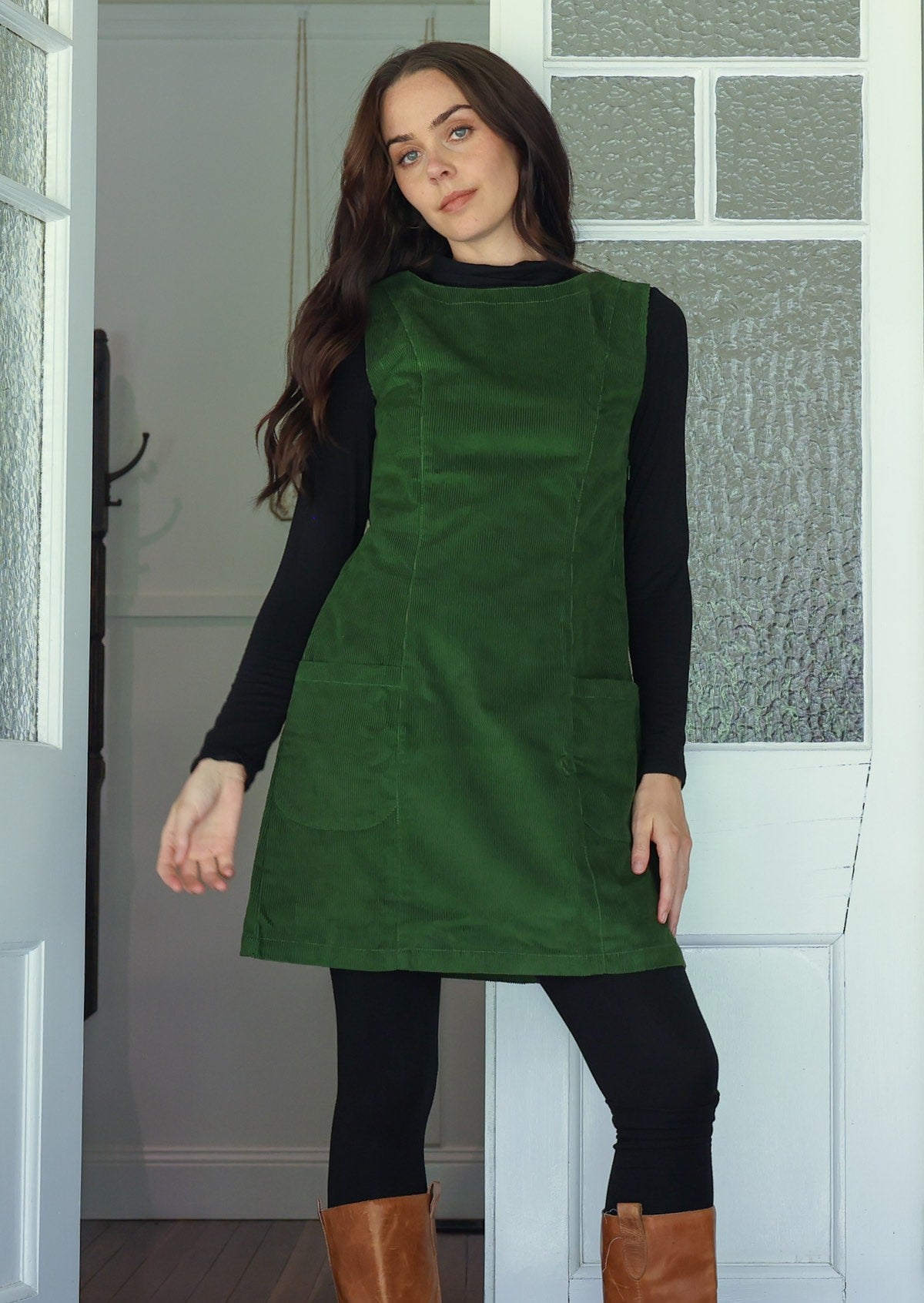 Woman wearing a green cotton corduroy sleeveless tunic layered over a black top and leggings and tan boots
