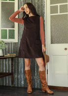 Woman in a dark brown sleeveless dress with boat neck and brown boots standing in front of a door with textured glass panel