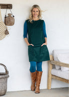 Woman wearing a dark green sleeveless cotton tunic over a teal long-sleeve top and leggings, and brown knee-high boots, with hands in pockets