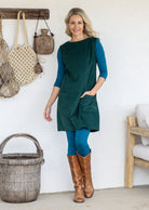 Woman wearing a sleeveless dark green cotton tunic over teal long-sleeve top and leggings and tan knee-high boots, with one hand in pocket