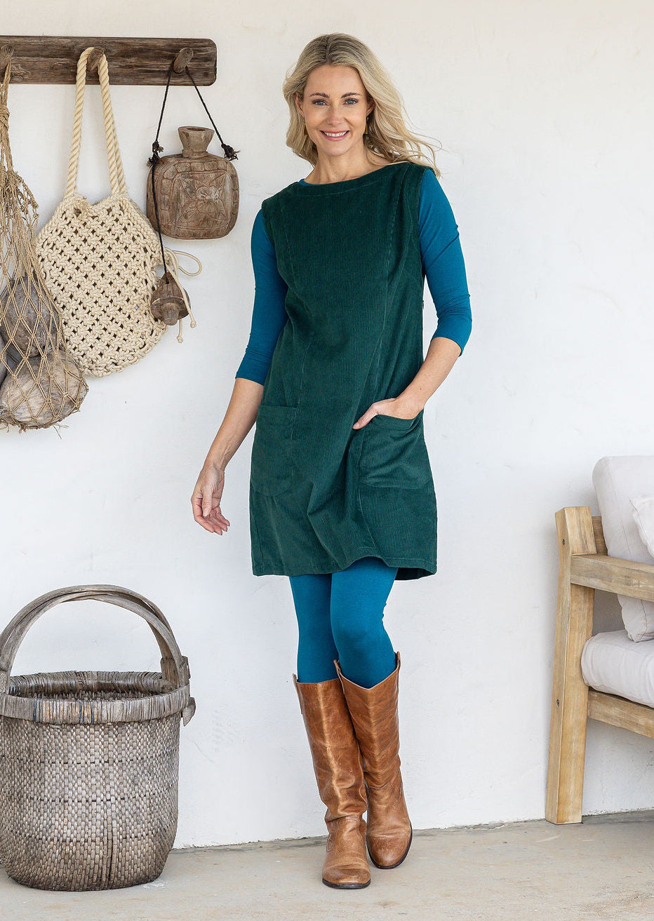 Woman wearing a sleeveless dark green cotton tunic over teal long-sleeve top and leggings and tan knee-high boots, with one hand in pocket