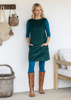 A woman wearing a dark green sleeveless cotton pinafore dress over a teal long-sleeve top and leggings, paired with brown knee-high boots, with hands in pockets