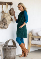 A woman wearing a dark green cotton corduroy tunic over a teal long-sleeve top and leggings, standing in a rustic room with a woven basket, wooden furniture